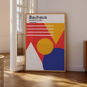 Puede incluir: Un póster rectangular enmarcado con el texto Bauhaus AUSSTELLUNG en la parte superior. El diseño incluye un círculo amarillo grande, un círculo azul, un triángulo blanco y un triángulo amarillo sobre un fondo rojo con líneas horizontales blancas.