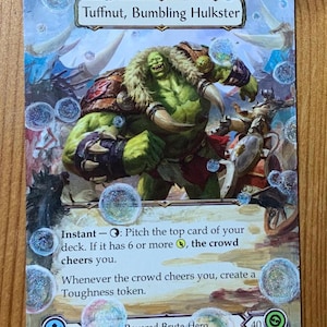 May include: A trading card featuring "Tuffnut, Bumbling Hulkster." The card depicts a green-skinned, muscular character with a fur collar. Text includes "Instant" and "Revered Brute Hero." The card has a blue and white color scheme with a 40 life point value.
