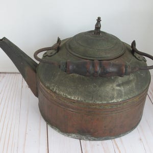 May include: A vintage kettle featuring a weathered copper body with a tarnished metallic top and lid. It includes a dark brown wooden handle attached to a metal bail, a long angled spout, and a small decorative finial on the center of the lid.