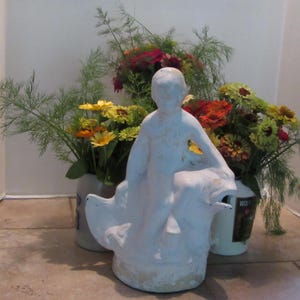 May include: White ceramic figurine of a person riding a fish. The figurine is surrounded by yellow, orange, and red flowers in white ceramic pots.
