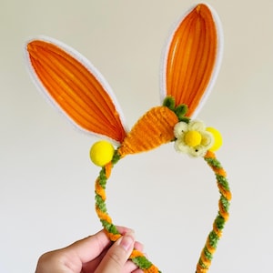 May include: A festive headband featuring orange bunny ears with white trim, a carrot, and a flower. The headband is decorated with yellow pom-poms and a green and orange twisted design, perfect for Easter celebrations.
