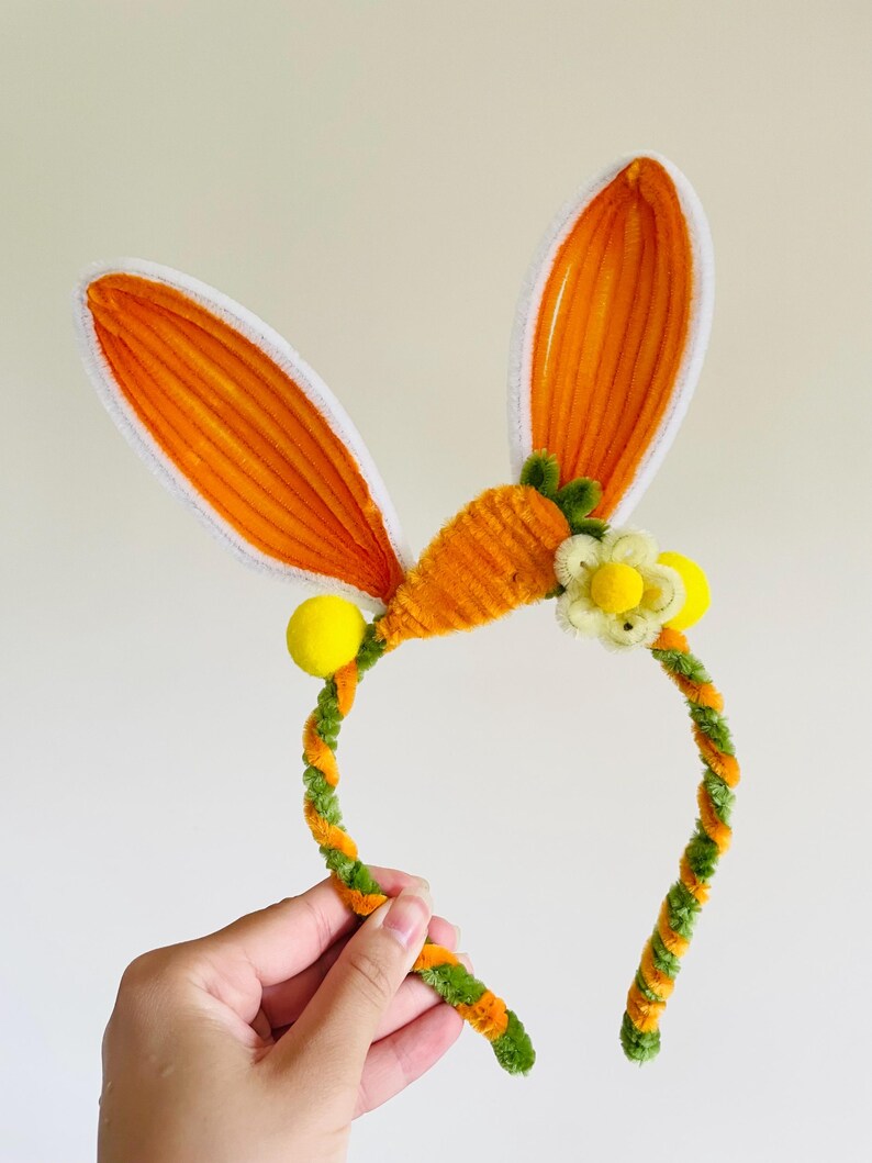 May include: A festive headband featuring orange bunny ears with white trim, a carrot, and a flower. The headband is decorated with yellow pom-poms and a green and orange twisted design, perfect for Easter celebrations.