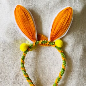 May include: A festive Easter headband featuring orange bunny ears with white trim. The headband is decorated with green and orange twisted pipe cleaners and yellow pom-poms, creating a playful and colorful accessory.