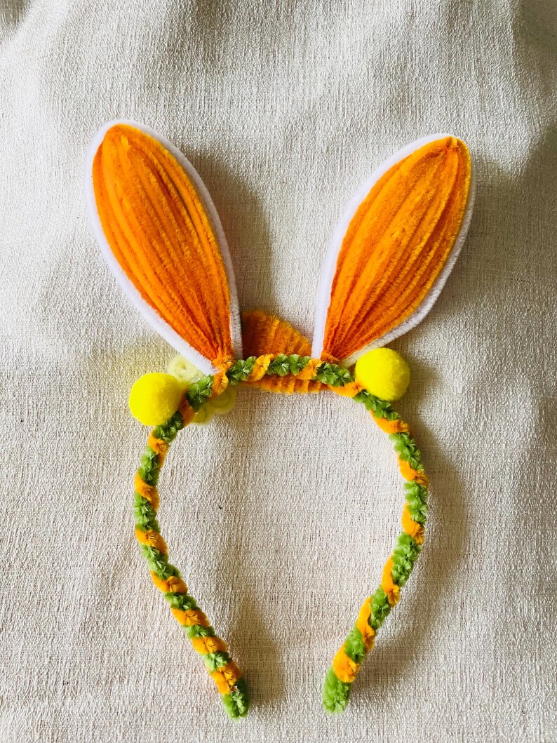 May include: A festive Easter headband featuring orange bunny ears with white trim. The headband is decorated with green and orange twisted pipe cleaners and yellow pom-poms, creating a playful and colorful accessory.
