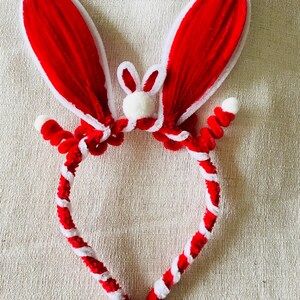 May include: A red and white bunny ear headband. The ears are red with white trim, and the headband is red and white twisted. A small white bunny face is centered between the ears. This festive accessory is perfect for Easter or themed events.