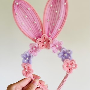 May include: A pink bunny ear headband with faux pearls and floral accents. The ears are a light pink with white faux pearls. The headband features pink and purple flowers.