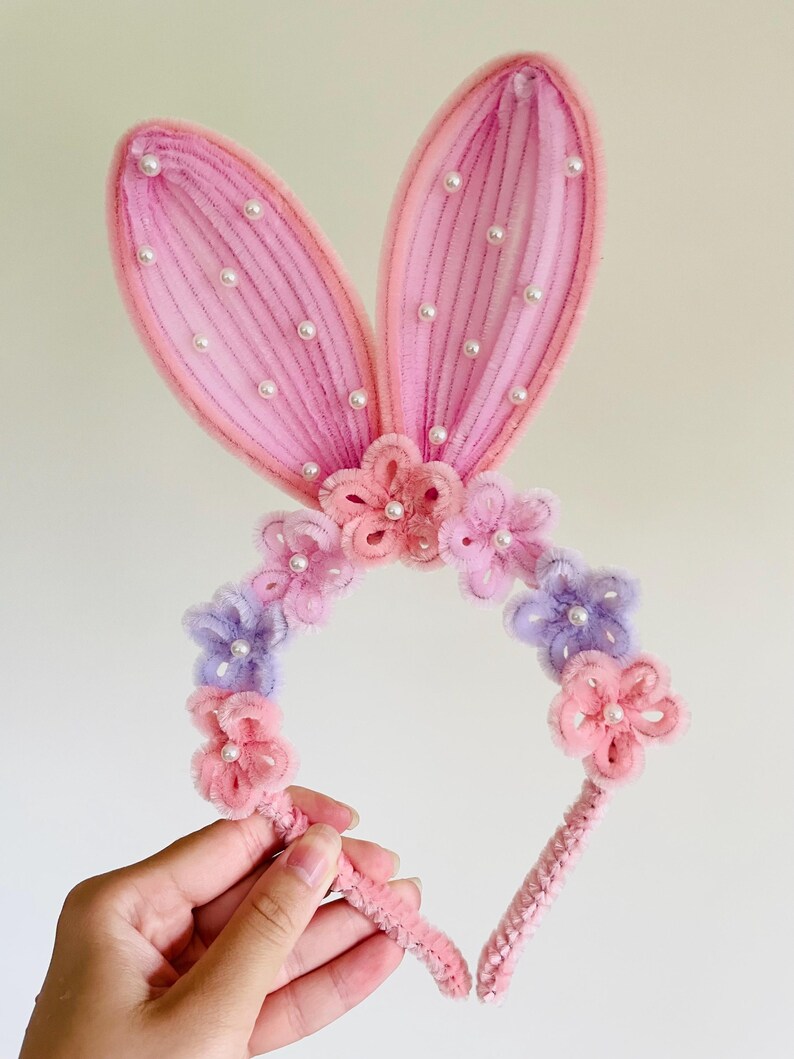 May include: A pink bunny ear headband with faux pearls and floral accents. The ears are a light pink with white faux pearls. The headband features pink and purple flowers.