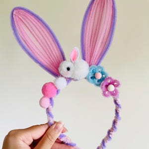 May include: A whimsical Easter bunny ear headband. The ears are pink with purple trim, and the headband is adorned with pom-poms and flowers. A fluffy white bunny is centered between the ears. The headband is held by a hand.