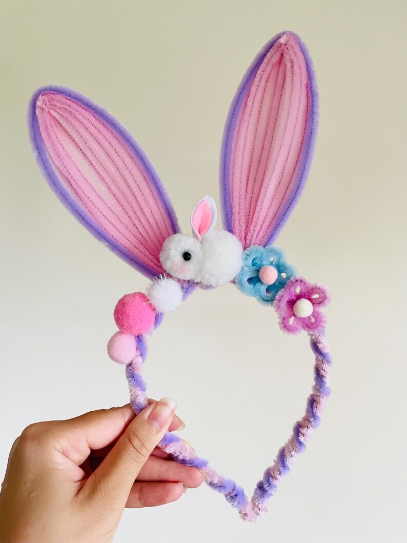 May include: A whimsical Easter bunny ear headband. The ears are pink with purple trim, and the headband is adorned with pom-poms and flowers. A fluffy white bunny is centered between the ears. The headband is held by a hand.