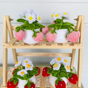 May include: Four small white ceramic pots with handmade strawberry and daisy floral arrangements. The strawberries are pink or red with white dots, and the daisies have white petals and yellow centres. The pots sit on a wooden shelf.