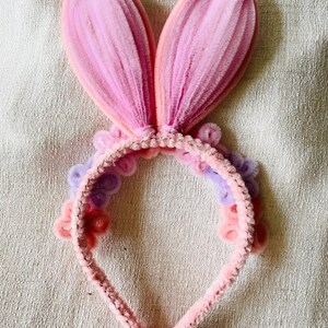 May include: A pink bunny ear headband with a soft, textured appearance. The ears are a gradient of pink hues, and the headband is adorned with small, decorative flowers in shades of pink and purple. A festive accessory.