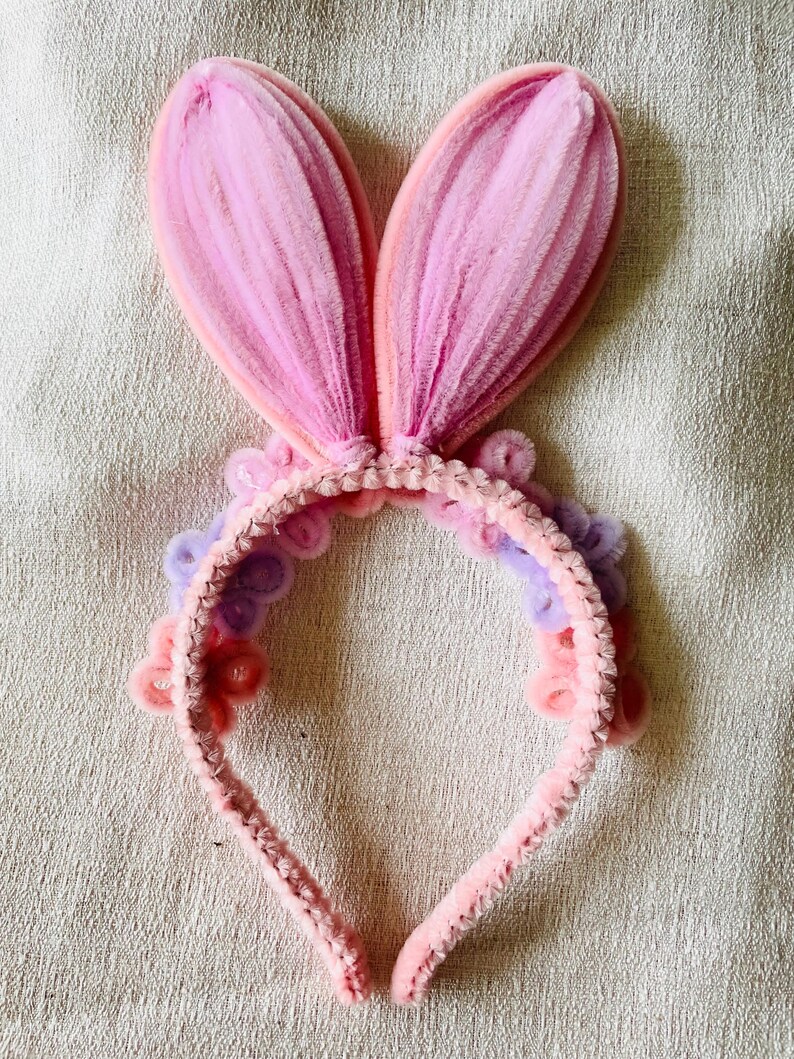 May include: A pink bunny ear headband with a soft, textured appearance. The ears are a gradient of pink hues, and the headband is adorned with small, decorative flowers in shades of pink and purple. A festive accessory.