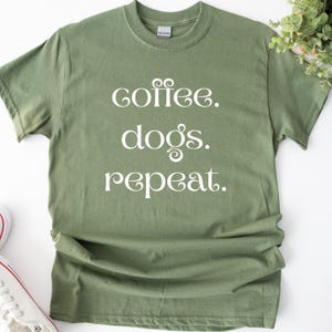 Könnte beinhalten: Olivgrünes T-Shirt mit dem weißen Schriftzug "coffee. dogs. repeat." in dekorativer Schrift. Das Shirt hat einen Rundhalsausschnitt und ist aus einem weichen Stoff gefertigt. Ein bequemes, lässiges Oberteil.