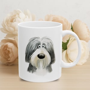 以下が含まれることがあります： 白いセラミックマグカップ。水彩画で描かれたバーデッドコリー犬のイラストが特徴です。犬は黒とグレーの毛並み、長い白いひげ、黒い鼻をしています。マグカップはカーブしたハンドル付きで、ソフトなクリーム色の花の背景に配置されています。