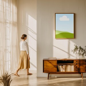 Puede incluir: Una impresi&oacute;n art&iacute;stica enmarcada de un paisaje con cielo azul, nube blanca y colinas verdes cuelga de una pared blanca. Debajo de la obra de arte hay un mueble de madera con libros y una planta. Una persona con una blusa blanca y una falda color canela pasa.