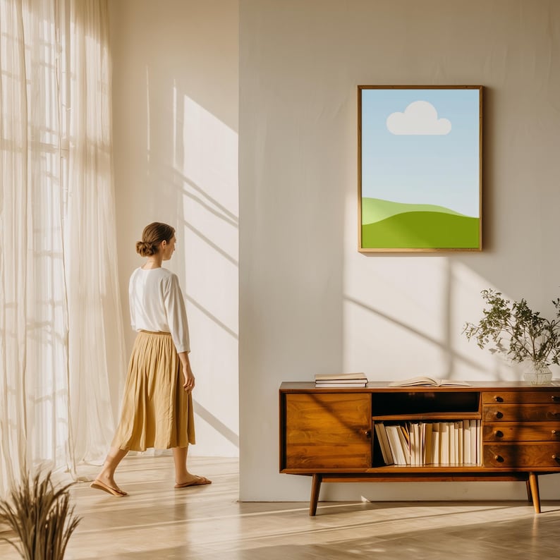 Puede incluir: Una impresi&oacute;n art&iacute;stica enmarcada de un paisaje con cielo azul, nube blanca y colinas verdes cuelga de una pared blanca. Debajo de la obra de arte hay un mueble de madera con libros y una planta. Una persona con una blusa blanca y una falda color canela pasa.