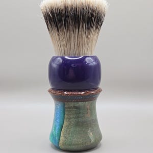 May include: A shaving brush with a dark purple handle and a two-tone green and blue base. The brush head is white with black tips. The brush is designed for grooming.