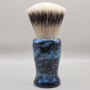 May include: A shaving brush with a blue and black marbled handle and a white and black bristle head. The brush has a rounded shape and is designed for applying shaving cream or soap to the face before shaving.