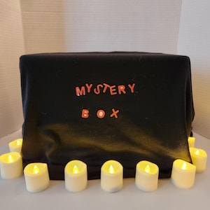 May include: A black box draped with black fabric, with the words "MYSTERY BOX" in red letters. The box is surrounded by several small, white, battery-operated candles with flickering yellow lights, creating a warm ambiance.