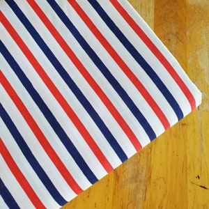 May include: White fabric with red and navy blue horizontal stripes. The fabric is folded in half and lying on a wooden surface.