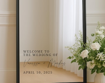 Pode incluir: Um espelho retangular grande com moldura preta e o texto "WELCOME TO THE WEDDING OF Vanessa + Markus APRIL 10, 2025" em escrita preta. Um vaso de flores brancas está à direita do espelho.