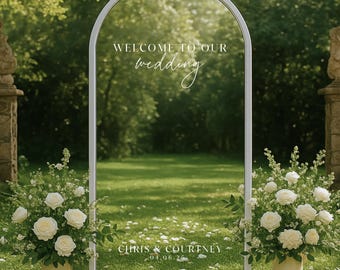 May include: A white arched wedding sign with the text "WELCOME TO OUR wedding" and names "CHRIS & COURTNEY" with a date. The sign is flanked by two arrangements of white roses in cream-colored vases, set on a green lawn.