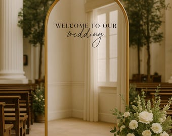 May include: A gold arched mirror with the text "WELCOME TO OUR wedding" and the names "ANNA & JAMES" with the date 02.16.25. The mirror is in a wedding venue with floral arrangements and seating.