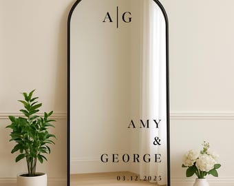 May include: A tall, arched black-framed mirror with the initials "A | G" at the top and the names "AMY & GEORGE" with the date "03.12.2025" in a black font. A potted plant is on the left and a vase of flowers on the right.