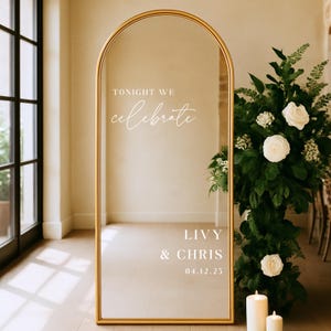 May include: A large, arched mirror with a gold frame. The mirror reflects the room and features the text "TONIGHT WE celebrate" at the top, with "LIVY & CHRIS 04.12.25" at the bottom. White roses and greenery are to the right.