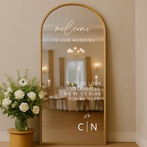 Può includere: Un grande specchio dorato ad arco con le parole "welcome to our wedding" in corsivo bianco. Sotto, il testo recita "You look incredible, we're so glad you're here!" e "xo C|N". Un'arrangiamento floreale è a sinistra.
