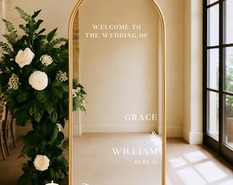 May include: A gold-framed, arched mirror with the text "WELCOME TO THE WEDDING OF GRACE & WILLIAM 05.03.25" and "Thank you for being here!". The mirror is placed in front of a floral arrangement and candles.
