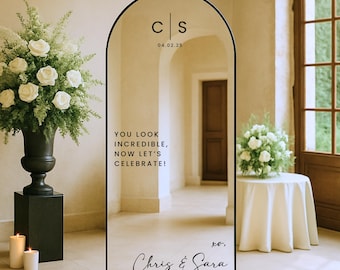 May include: A black arched mirror with the text "YOU LOOK INCREDIBLE, NOW LET'S CELEBRATE!" and the names "Chris & Sara". The mirror also displays the initials "CS" and the date "04.02.25". The mirror is a wedding or event decoration.