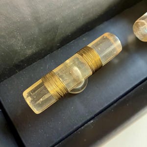May include: A clear, cylindrical glass bottle stopper with a gold-colored wire wrapped around the center. The stopper has a small, circular base and is set against a dark background.