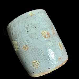 May include: A light blue ceramic mug with a hand-drawn face and grid pattern. The mug has a cylindrical shape with a slightly textured surface and is decorated with yellow and brown accents. The mug is set against a black background.