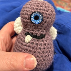 May include: A handmade crochet creature in shades of purple, featuring a large, detailed blue eye. It has small white wings and a black and white mouth detail. The amigurumi is held in a hand.