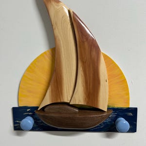 May include: Wooden sailboat wall decor with a yellow sun and blue water. The sailboat has light and dark wood sails and a brown hull. Two light blue knobs are below the boat, likely for hanging items. A nautical-themed decorative piece.