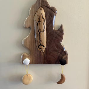 May include: A wooden wall hanging with a tree-like design, featuring light and dark brown wood. A central oval has a tree-like design. Two hanging ornaments include a white ball with a wooden disc and a black ball with a wooden crescent moon.