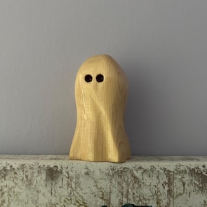 May include: A small, wooden ghost figurine with a smooth, light-colored finish. The ghost has two dark, round eyes and a simple, rounded shape. The figurine is a decorative item, likely for Halloween or spooky-themed decor.