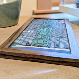 May include: A framed map print featuring a city grid with green and blue areas, likely representing parks and water. The print is set within a dark brown wooden frame, resting on a light-colored wooden surface. The map's details are visible.