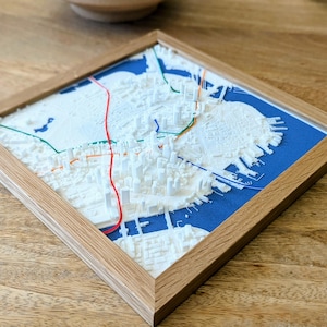 May include: A framed, three-dimensional white map of a city, set against a blue background. The map features raised structures and colorful lines, encased in a natural wood frame. The artwork is displayed on a wooden surface.