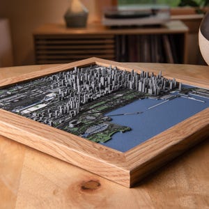 May include: A framed 3D map of a city, featuring a detailed depiction of buildings, parks, and a body of water. The map is set within a natural wood frame, resting on a wooden surface. The color palette includes shades of gray, green, and blue.