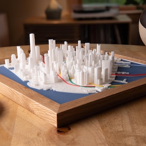 Può includere: Un modello 3D bianco di uno skyline cittadino, incorniciato in legno. Il modello si trova su una superficie blu, con linee rosse, verdi e gialle. Un sottobicchiere decorativo è in primo piano.