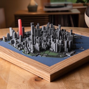 May include: A framed 3D model of a city skyline, likely New York City, with gray buildings and a prominent red tower. The model sits on a blue background within a wooden frame. The overall aesthetic is modern and architectural.