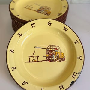 May include: A stack of yellow plates with a brown rim. Each plate features a brown illustration of a covered wagon and a campfire. Brown letters are spaced around the rim. The plates have a vintage, rustic aesthetic.