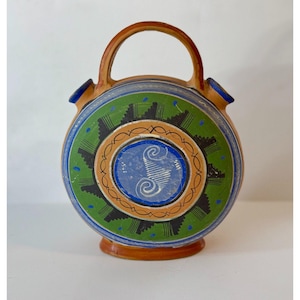 May include: A round, hand-painted ceramic flask with a handle. The flask features a central blue design with a white spiral, surrounded by green, black, and orange patterns. The rim and handle are brown, and the spout is blue.