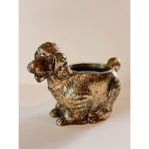 Puede incluir: Un recipiente plateado con forma de perro con una abertura en forma de cuenco. El perro está sentado, con textura de pelaje detallada y un collar. El recipiente es probablemente para uso decorativo o funcional.