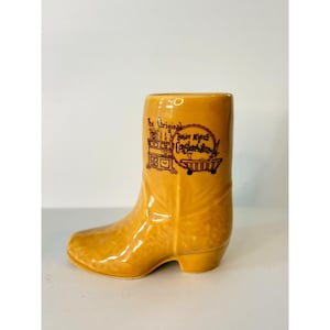 May include: A ceramic cowboy boot in a warm yellow color. The boot features a detailed design with the text "The Original Bobby Myers Conglomeration" in brown. The boot is a decorative item.