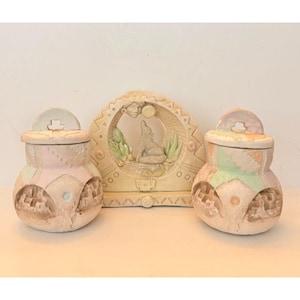 May include: A set of three ceramic decorative pieces. Two small jars with lids are on either side of a larger decorative arch. The jars are pastel pink and green with geometric patterns. The arch features a howling wolf and moon design.