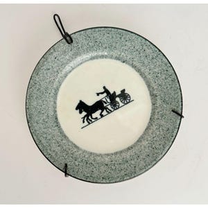 May include: A decorative plate with a speckled gray rim and a white center. The center features a black silhouette of a horse-drawn carriage. The plate has a black wire hanger attached for display.
