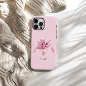 May include: A pale pink phone case with a pink lily flower design. The case has the name "MAEVE" printed beneath the flower. The phone case is displayed on a white surface with a cream-coloured knitted sweater in the background.